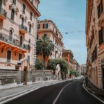 Strada curva a Roma fiancheggiata da palazzi storici color ocra e rosa, balconi in ferro battuto e una palma; cielo sereno, insegna “Sementi Piante” a destra, nessun traffico.