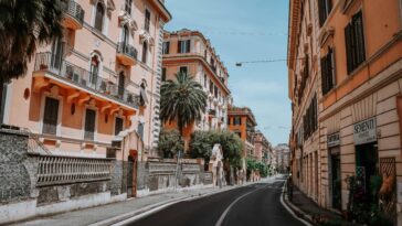 Strada curva a Roma fiancheggiata da palazzi storici color ocra e rosa, balconi in ferro battuto e una palma; cielo sereno, insegna “Sementi Piante” a destra, nessun traffico.