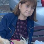 Giuseppina Giuliano donna sui 60 anni seduta su una panchina, giacca blu, abito scuro a pois, con un piccolo chihuahua chiaro in grembo; auto e sanpietrini sullo sfondo.