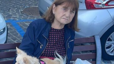 Giuseppina Giuliano donna sui 60 anni seduta su una panchina, giacca blu, abito scuro a pois, con un piccolo chihuahua chiaro in grembo; auto e sanpietrini sullo sfondo.