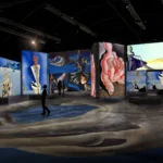 Spazio espositivo buio con grandi schermi che proiettano opere surrealiste; visitatori in controluce camminano tra installazioni e proiezioni.