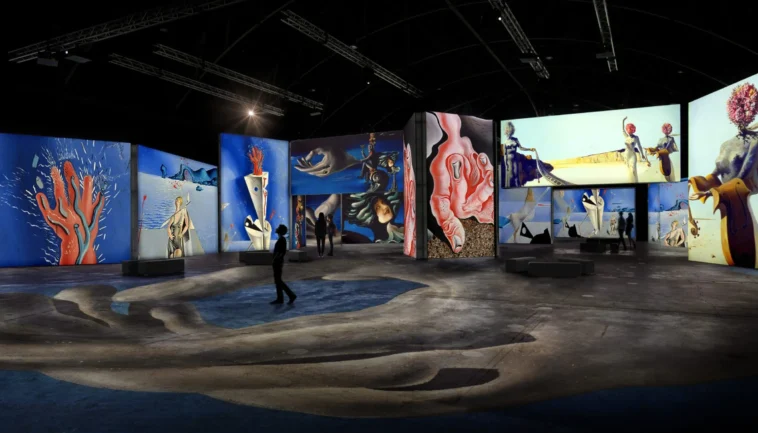 Spazio espositivo buio con grandi schermi che proiettano opere surrealiste; visitatori in controluce camminano tra installazioni e proiezioni.