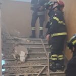 Vigili del Fuoco su una scala coperta di macerie dopo il crollo del controsoffitto in una scuola; polvere nell’aria e corrimano danneggiati.