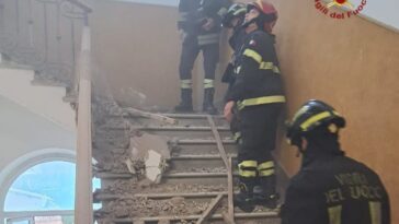 Vigili del Fuoco su una scala coperta di macerie dopo il crollo del controsoffitto in una scuola; polvere nell’aria e corrimano danneggiati.