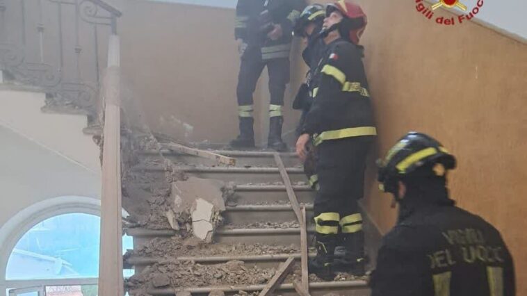 Vigili del Fuoco su una scala coperta di macerie dopo il crollo del controsoffitto in una scuola; polvere nell’aria e corrimano danneggiati.