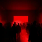 Persone in controluce camminano verso uno schermo rosso acceso in una sala buia, avvolta da fumo e luci intense durante un evento.