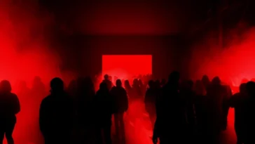 Persone in controluce camminano verso uno schermo rosso acceso in una sala buia, avvolta da fumo e luci intense durante un evento.