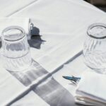Tavolo con tovaglia bianca, due bicchieri capovolti e tovaglioli con posate; luce intensa e ombre nette, atmosfera da ristorante all’aperto.