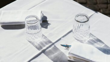 Tavolo con tovaglia bianca, due bicchieri capovolti e tovaglioli con posate; luce intensa e ombre nette, atmosfera da ristorante all’aperto.