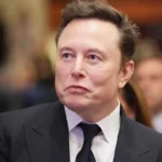 Primo piano di un uomo con giacca e cravatta nera, volto rivolto di lato; sullo sfondo persone sfocate in una sala conferenze.