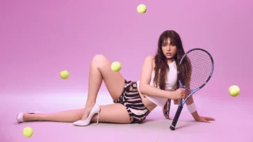 Modella su sfondo rosa, sdraiata con racchetta da tennis in mano, palline gialle sospese intorno; top bianco, shorts fantasia, décolleté bianche.
