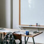 Aula scolastica con banchi in legno, zaini e quaderni, grande lavagna bianca su parete e luce da finestra.