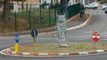 Piramide di carrelli della spesa impilati al centro di una rotatoria in via del Torraccio, Villa Verde (Roma), con cartelli stradali attorno.