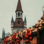 Decorazioni natalizie rosse e lucine bokeh in primo piano con torre campanaria in stile gotico sullo sfondo, scena tipica dei Mercatini di Natale.