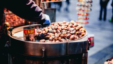 Un venditore prepara caldarroste su un grande braciere: il prezzo segnalato è 5,00 €. Scene tipiche del weekend di Tutti i Santi, tra mercatini e passeggiate nei quartieri della Capitale.