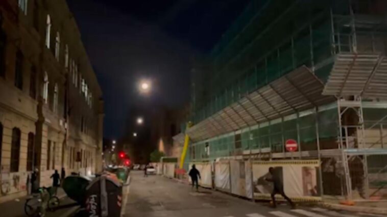 Via Campania di notte, con il liceo Righi a sinistra e un edificio coperto da impalcature a destra; alcuni ragazzi incappucciati corrono vicino alle transenne del cantiere.