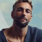 Uomo barbuto con maglione scuro in primo piano, sfondo chiaro.