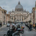 Veduta di via della Conciliazione a Roma con scooter e auto in movimento e la basilica di San Pietro sullo sfondo in una giornata nuvolosa.