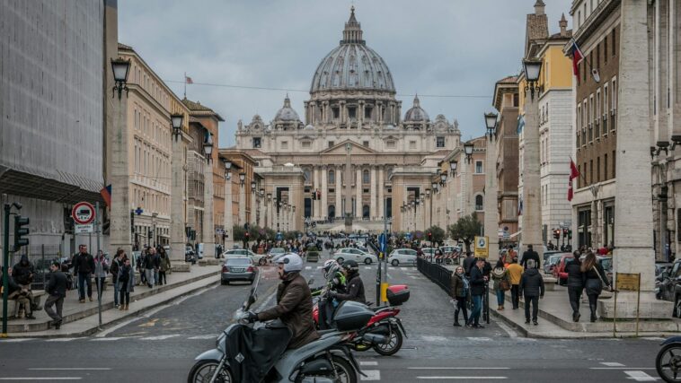Veduta di via della Conciliazione a Roma con scooter e auto in movimento e la basilica di San Pietro sullo sfondo in una giornata nuvolosa.
