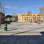 Veduta di piazza Erasmo Piaggio a Villaggio Breda, con palazzi residenziali, alberi spogli invernali, area giochi e persone che attraversano la piazza.