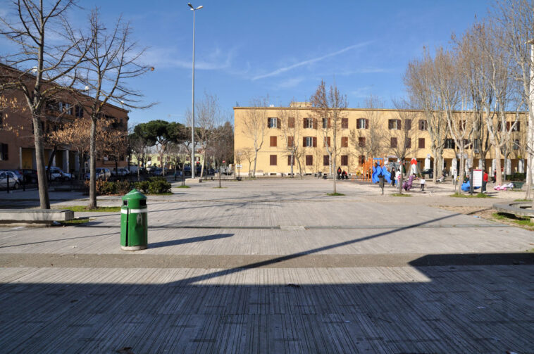 Veduta di piazza Erasmo Piaggio a Villaggio Breda, con palazzi residenziali, alberi spogli invernali, area giochi e persone che attraversano la piazza.