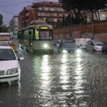 Tram e auto attraversano una strada completamente allagata a Roma durante un forte temporale, con pioggia intensa e traffico rallentato.