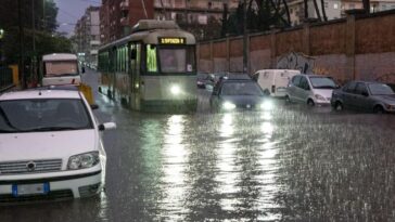 Tram e auto attraversano una strada completamente allagata a Roma durante un forte temporale, con pioggia intensa e traffico rallentato.