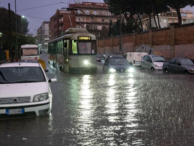 Tram e auto attraversano una strada completamente allagata a Roma durante un forte temporale, con pioggia intensa e traffico rallentato.