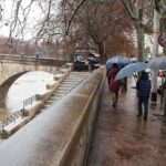 Persone con ombrelli camminano sul lungotevere bagnato durante una giornata di pioggia a Roma, con il fiume ingrossato e un ponte storico sullo sfondo.