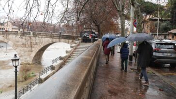 Persone con ombrelli camminano sul lungotevere bagnato durante una giornata di pioggia a Roma, con il fiume ingrossato e un ponte storico sullo sfondo.