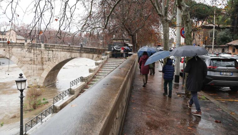 Persone con ombrelli camminano sul lungotevere bagnato durante una giornata di pioggia a Roma, con il fiume ingrossato e un ponte storico sullo sfondo.