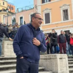 Carlo Conti sorride sulla scalinata di Piazza di Spagna circondato da fan e curiosi durante le riprese dello spot “Tutti cantano Sanremo”.