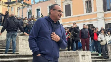 Carlo Conti sorride sulla scalinata di Piazza di Spagna circondato da fan e curiosi durante le riprese dello spot “Tutti cantano Sanremo”.