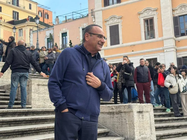 Carlo Conti sorride sulla scalinata di Piazza di Spagna circondato da fan e curiosi durante le riprese dello spot “Tutti cantano Sanremo”.