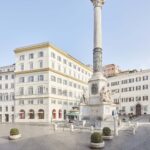 Vista di piazza Mignanelli a Roma con la Colonna dell’Immacolata e gli edifici storici circostanti in una giornata luminosa.