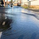 Strada completamente allagata su via Prenestina dopo la rottura di una tubatura, con auto e pedoni che cercano di avanzare tra l’acqua.