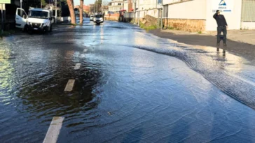 Strada completamente allagata su via Prenestina dopo la rottura di una tubatura, con auto e pedoni che cercano di avanzare tra l’acqua.