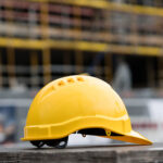 Casco da cantiere giallo appoggiato su una superficie di legno con un edificio in costruzione sullo sfondo.