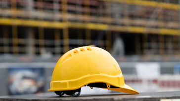 Casco da cantiere giallo appoggiato su una superficie di legno con un edificio in costruzione sullo sfondo.