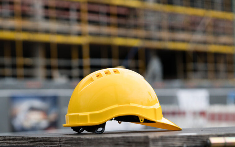 Casco da cantiere giallo appoggiato su una superficie di legno con un edificio in costruzione sullo sfondo.