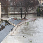 Banchine del Tevere allagate a Roma, con una persona sotto la pioggia e l’acqua del fiume a ridosso degli argini.