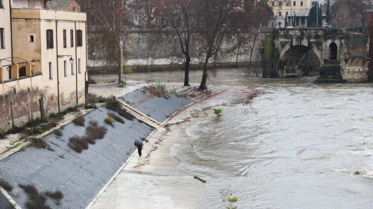 Banchine del Tevere allagate a Roma, con una persona sotto la pioggia e l’acqua del fiume a ridosso degli argini.