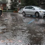 Primo piano di una strada di Roma completamente bagnata e parzialmente allagata dalla pioggia, con auto in transito e foglie trascinate dall’acqua.
