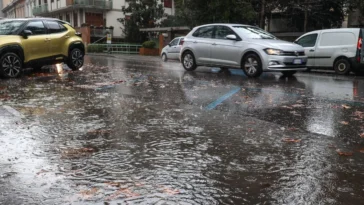 Primo piano di una strada di Roma completamente bagnata e parzialmente allagata dalla pioggia, con auto in transito e foglie trascinate dall’acqua.