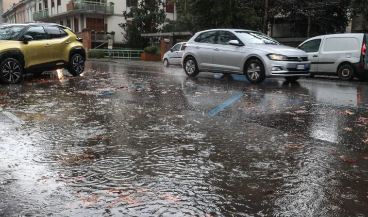 Primo piano di una strada di Roma completamente bagnata e parzialmente allagata dalla pioggia, con auto in transito e foglie trascinate dall’acqua.