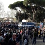 Folla radunata in strada a Roma durante un corteo, con diversi mezzi della polizia e agenti in tenuta antisommossa schierati per gestire la manifestazione.