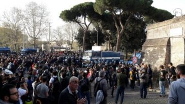 Folla radunata in strada a Roma durante un corteo, con diversi mezzi della polizia e agenti in tenuta antisommossa schierati per gestire la manifestazione.
