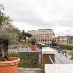 Vista dal belvedere Antonio Cederna con piante in vaso in primo piano e il Colosseo sullo sfondo, mentre pedoni percorrono via dei Fori Imperiali.