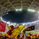 Tifosi della Roma allo Stadio Olimpico con bandiere giallorosse durante una partita serale.