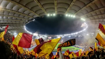 Tifosi della Roma allo Stadio Olimpico con bandiere giallorosse durante una partita serale.